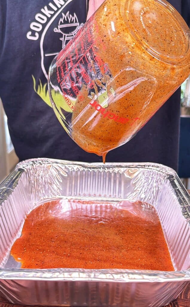 A person pours seasoned sauce from a glass measuring cup into a foil pan.