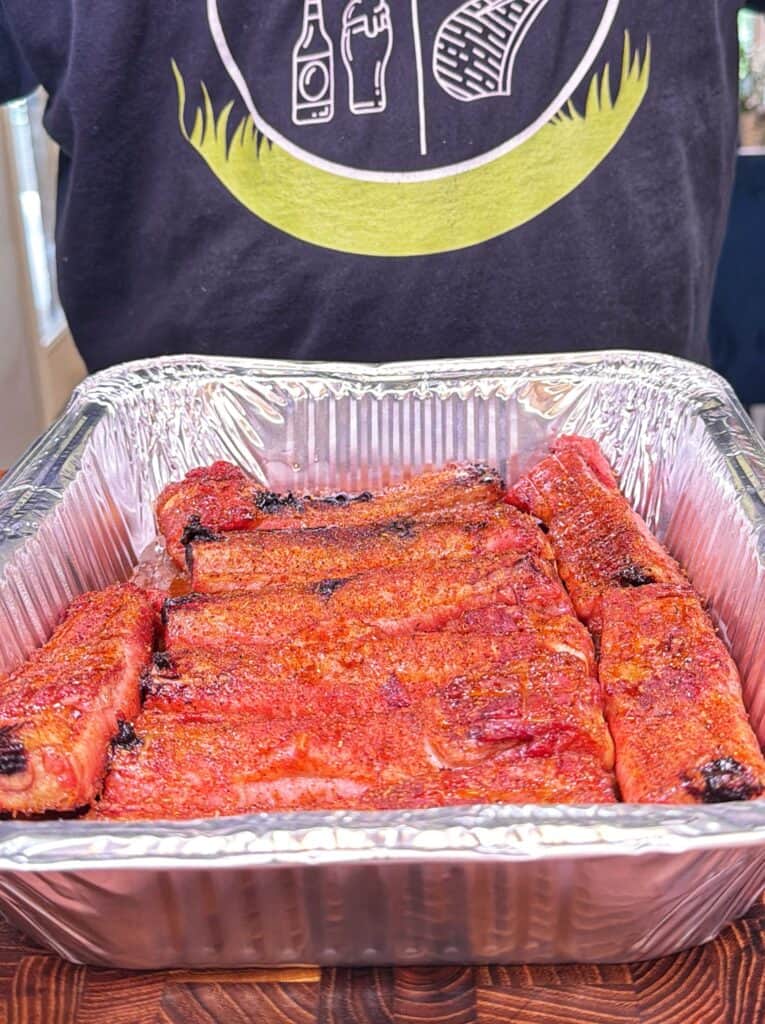 A person stands behind a foil pan filled with cooked seasoned pork ribs.
