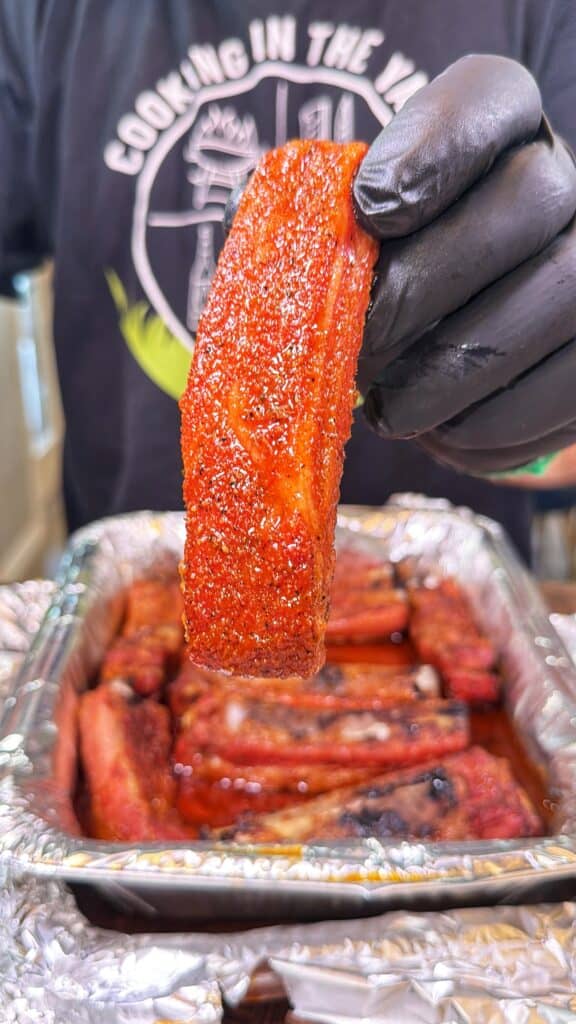A gloved hand holds a cooked seasoned pork rib over a foil-lined pan filled with ribs.