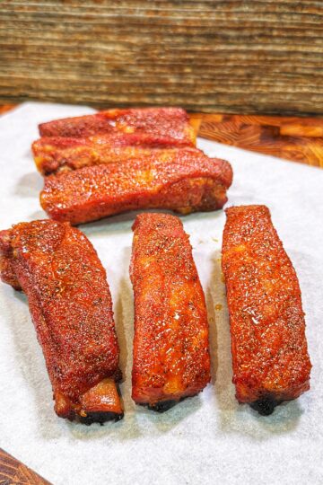 Five seasoned cooked pork ribs arranged on white parchment paper on a wooden surface.