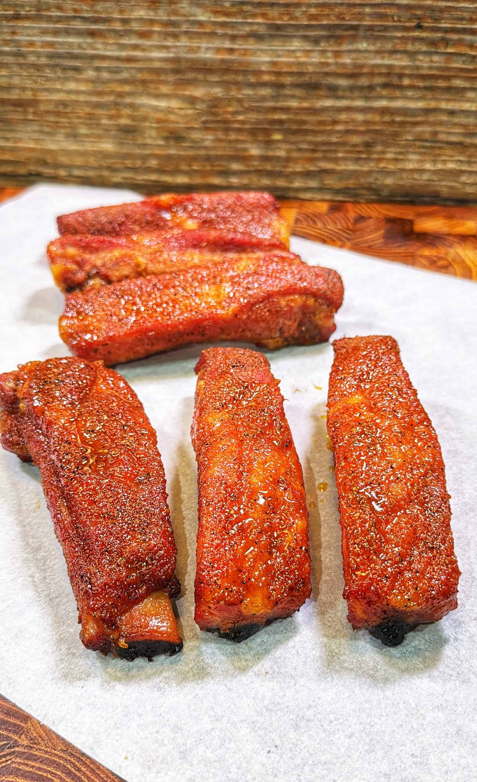 Five seasoned cooked pork ribs arranged on white parchment paper on a wooden surface.