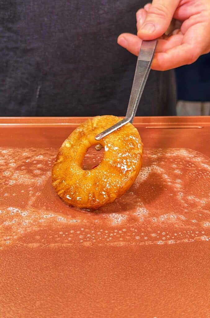 Metal tongs hold a pineapple ring over a hot griddle.