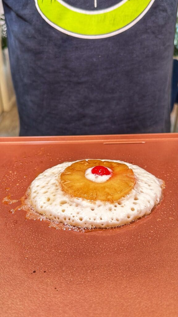 A pineapple ring with a cherry in the center sits on a pancake cooking on a griddle.