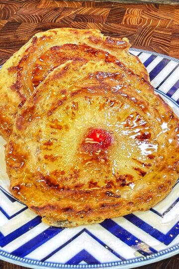 Two pineapple upside-down pancakes with cherries in the centers on a blue and white zigzag-patterned plate, with syrup visible.