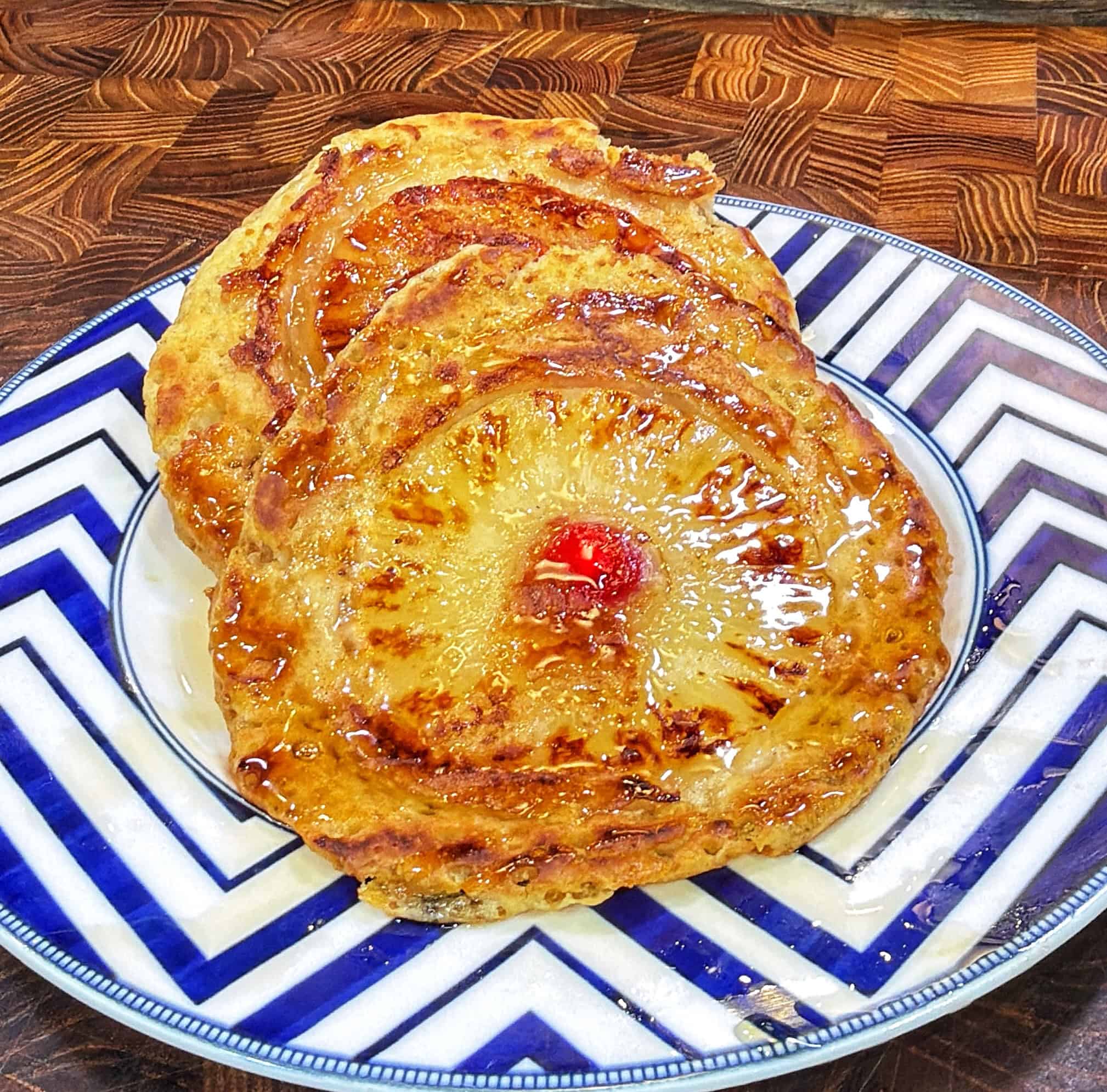 Two pineapple upside-down pancakes with cherries in the centers on a blue and white zigzag-patterned plate, with syrup visible.