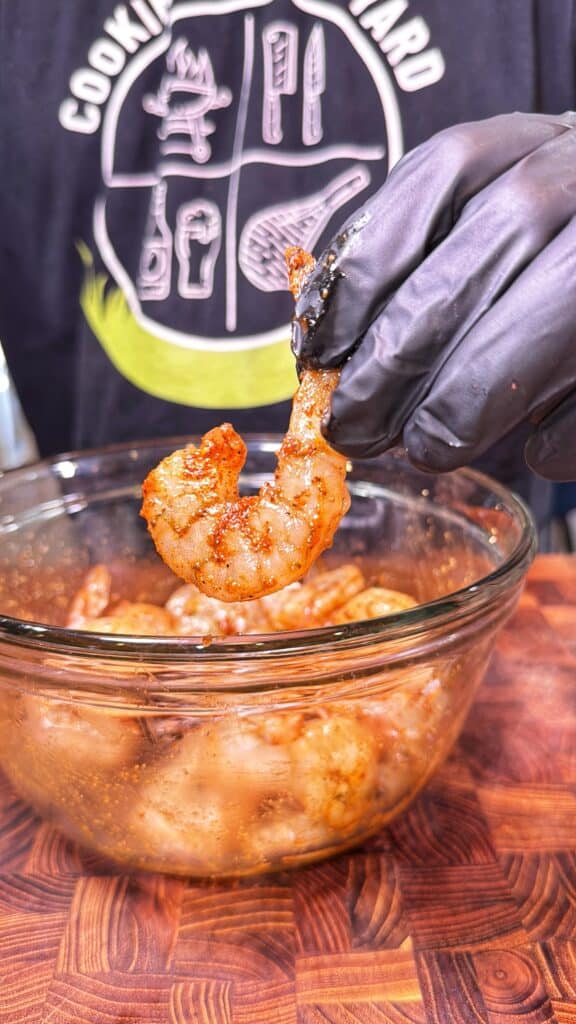 A gloved hand holds a seasoned shrimp over a bowl of shrimp.