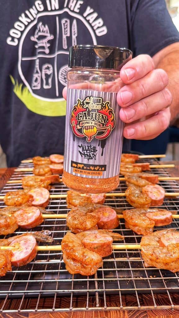 A jar of Cajun honey rub is held over sliced sausage on a wire rack.