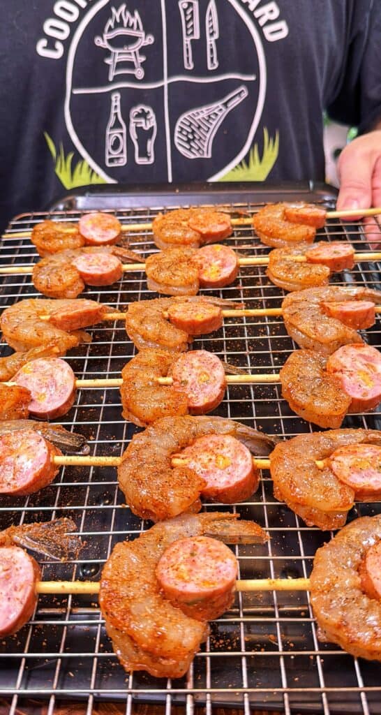 Seasoned shrimp and sausage are threaded onto skewers on a wire rack.