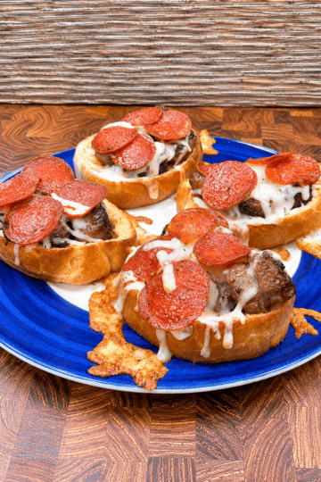 Four slices of bread topped with melted cheese, sausage, and pepperoni are arranged on a blue plate, all placed on a wooden surface.