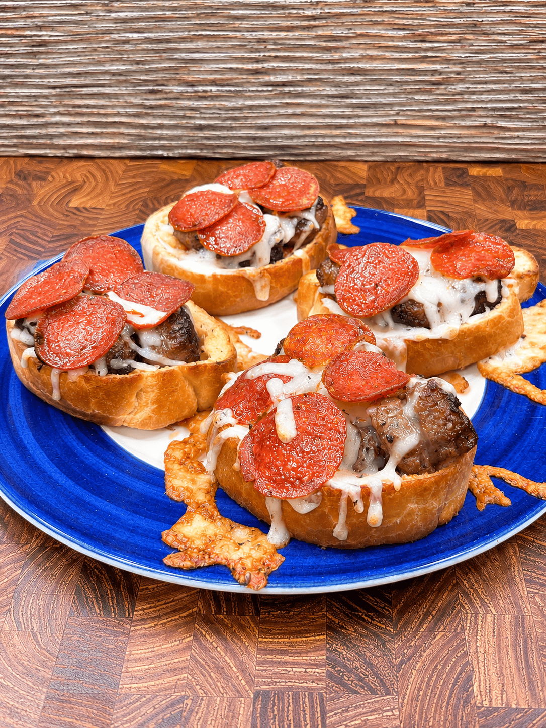 Four slices of bread topped with melted cheese, sausage, and pepperoni are arranged on a blue plate, all placed on a wooden surface.