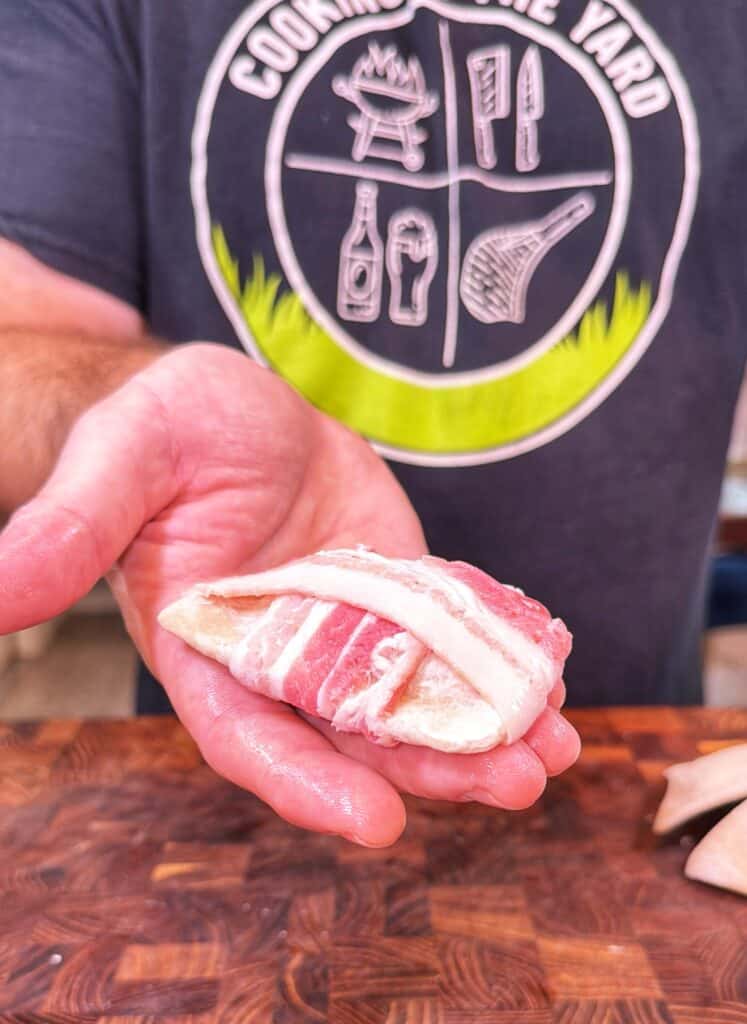 A person holds a raw bacon-wrapped pierogi over a wooden surface.