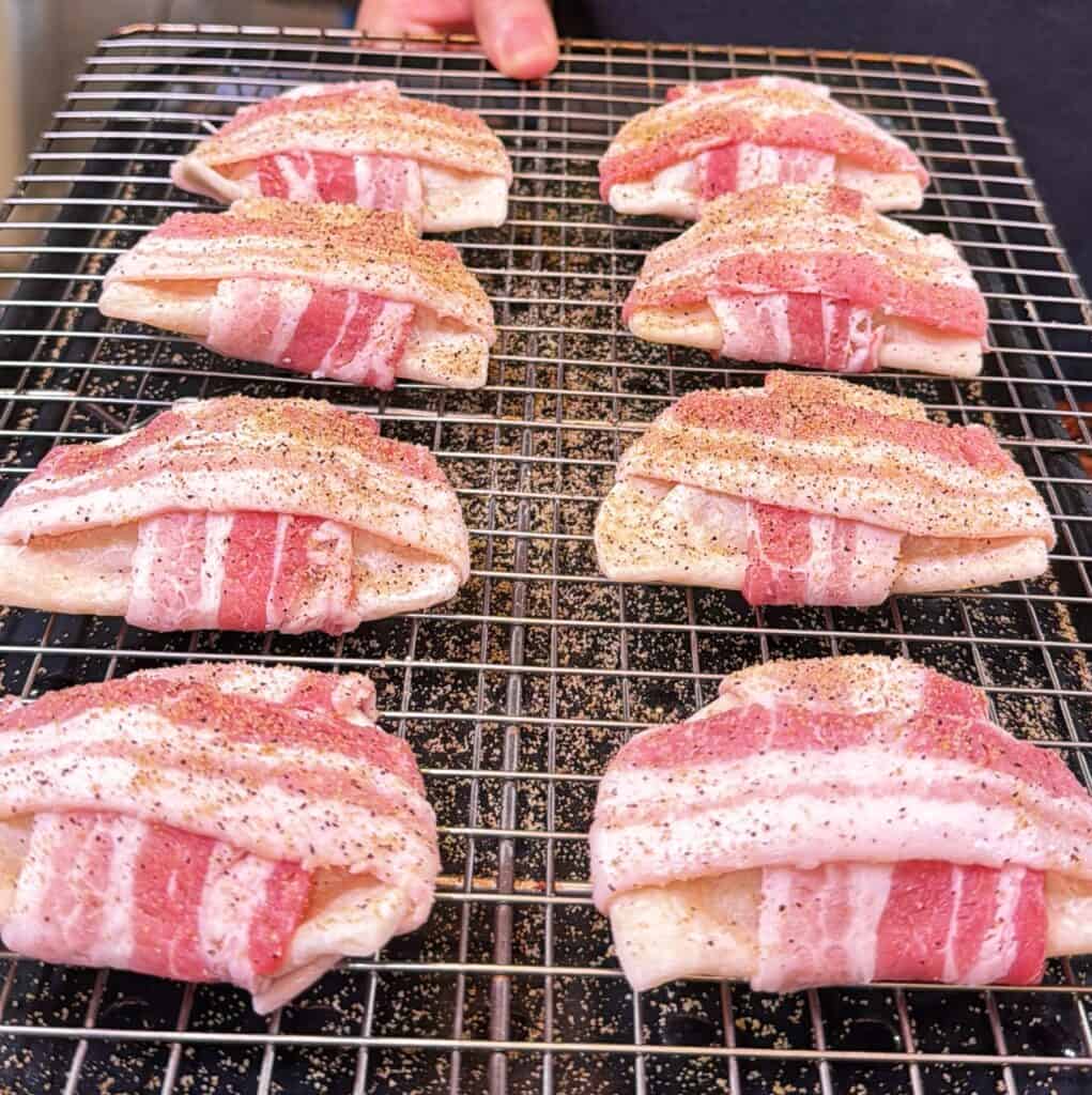 Raw bacon-wrapped pierogies seasoned with pepper are arranged in two rows on a metal cooling rack.