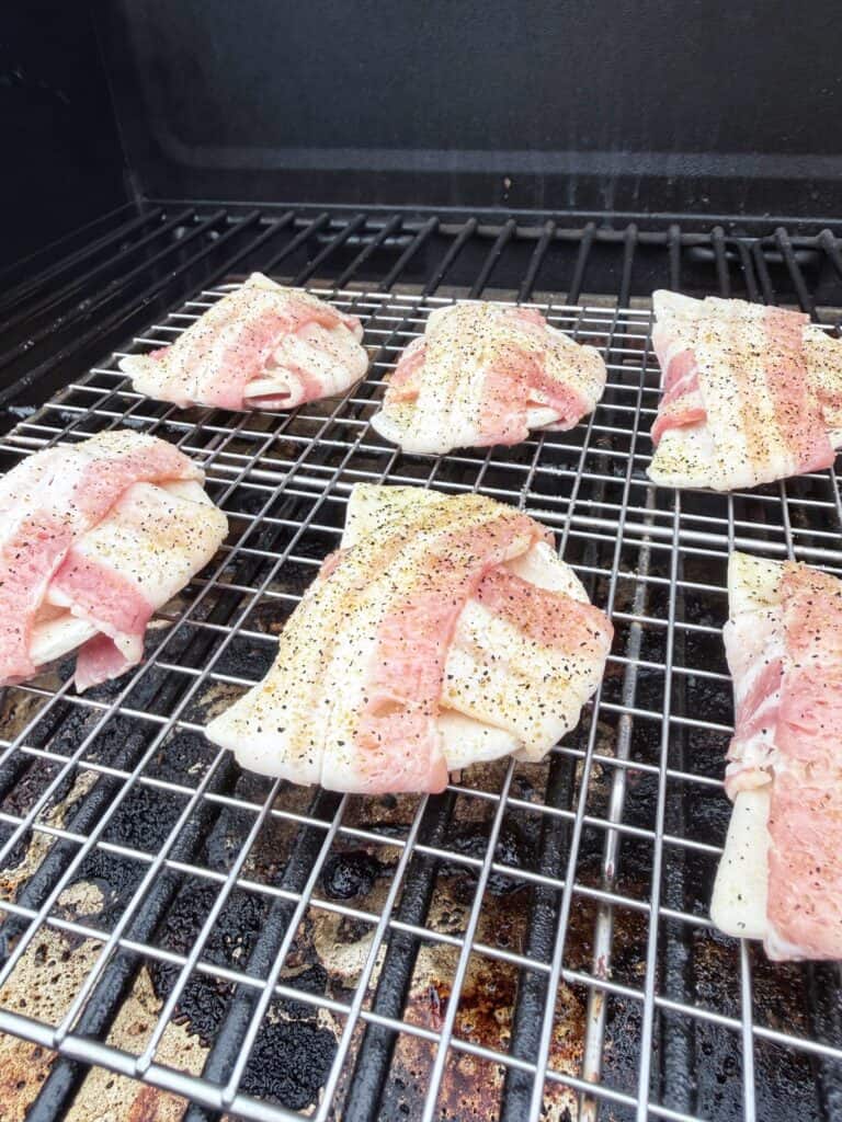 Bacon-wrapped pierogies seasoned with pepper cook on a metal grill inside a barbecue.