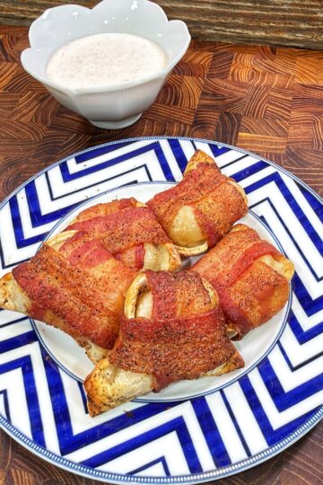 Four bacon-wrapped pierogies on a blue and white chevron-patterned plate, with a bowl of creamy dipping sauce in the background.