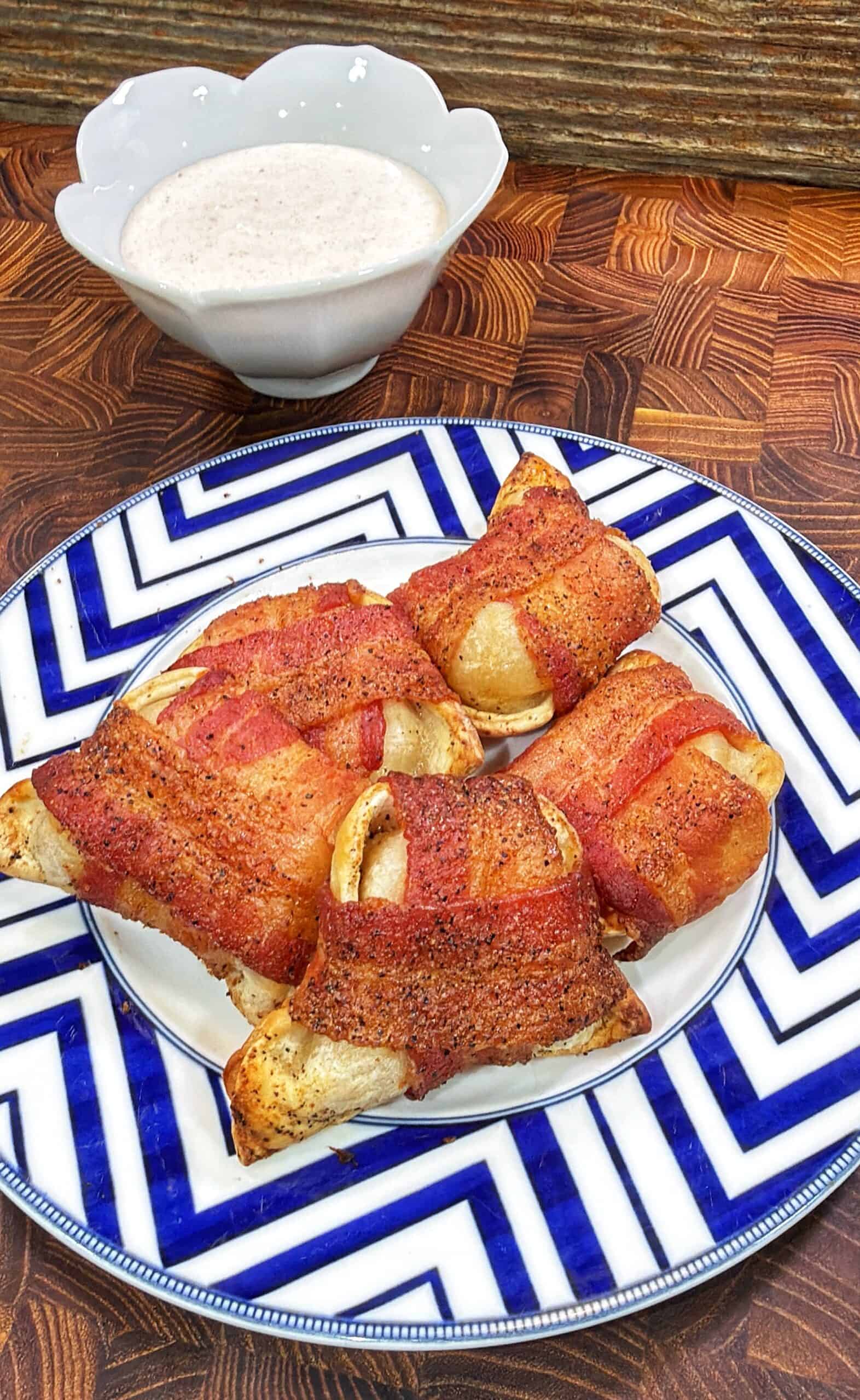 Four bacon-wrapped pierogies on a blue and white chevron-patterned plate, with a bowl of creamy dipping sauce in the background.