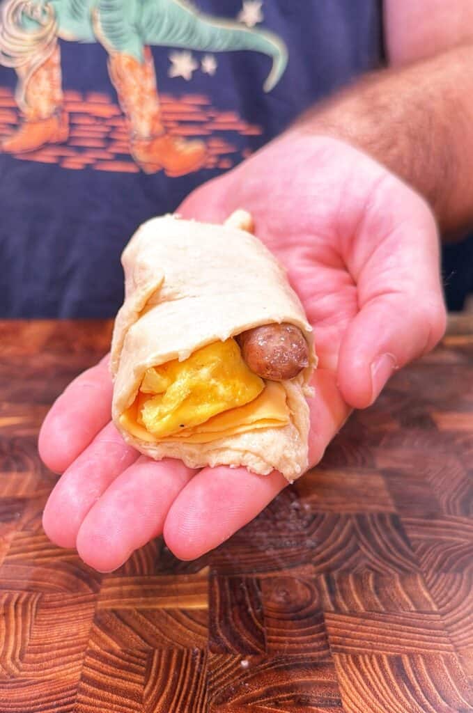 A hand holds a crescent roll-up filled with scrambled eggs, cheese, and breakfast sausage.
