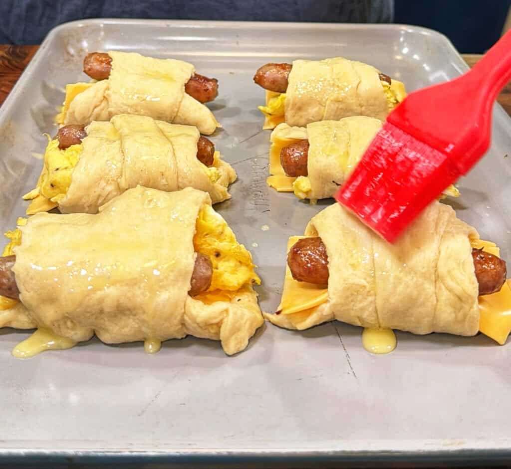 A hand brushes egg wash over unbaked sausage, egg, and cheese crescent roll-ups on a baking sheet.