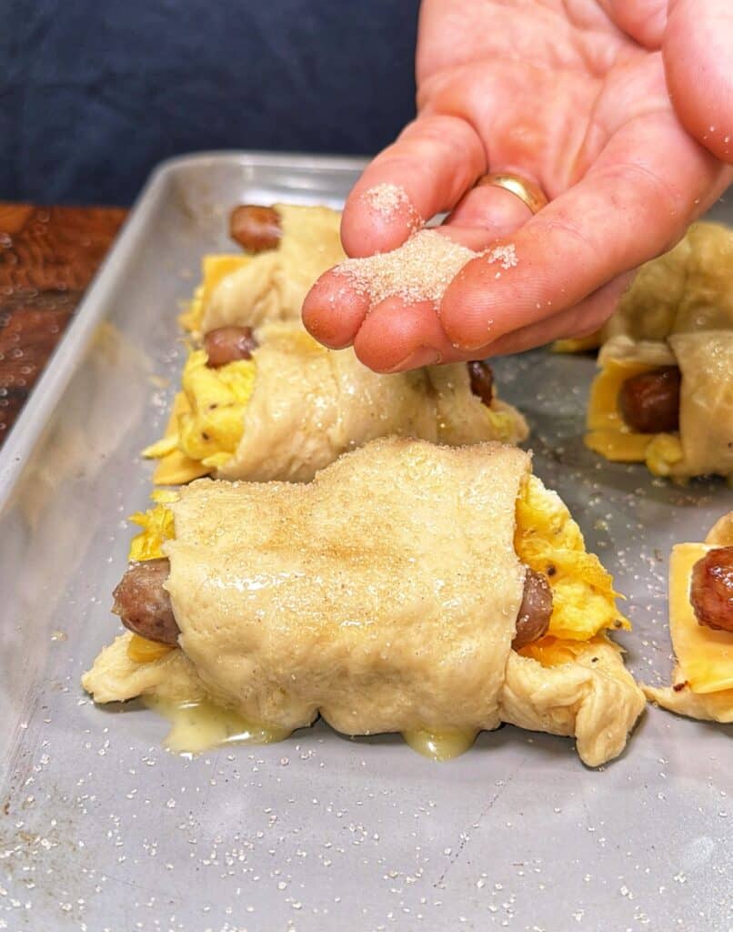 A hand sprinkles cinnamon sugar over unbaked sausage, egg, and cheese crescent roll-ups on a baking tray.