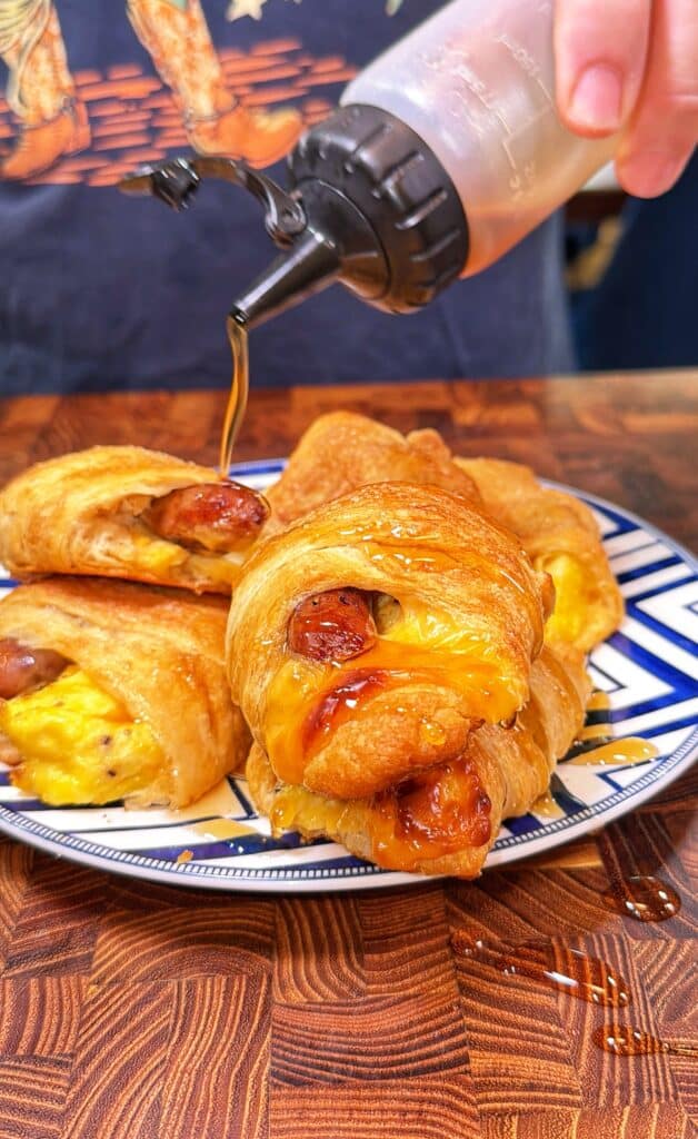 A hand pours maple syrup over sausage, egg, and cheese crescent roll-ups on a plate.