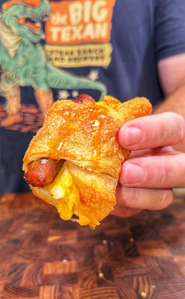 A hand holds a crescent roll-up wrapped around sausage and melted cheese.