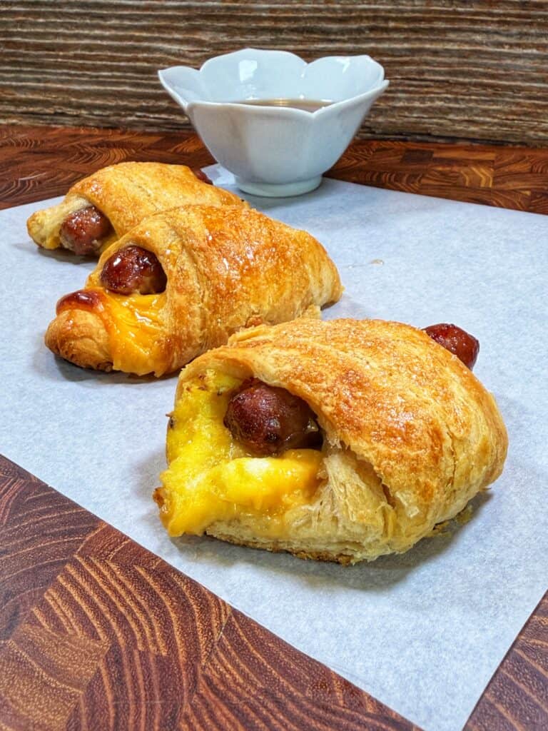 Two sausage, egg, and cheese crescent roll-ups sit on parchment paper with a small bowl of dipping sauce nearby.