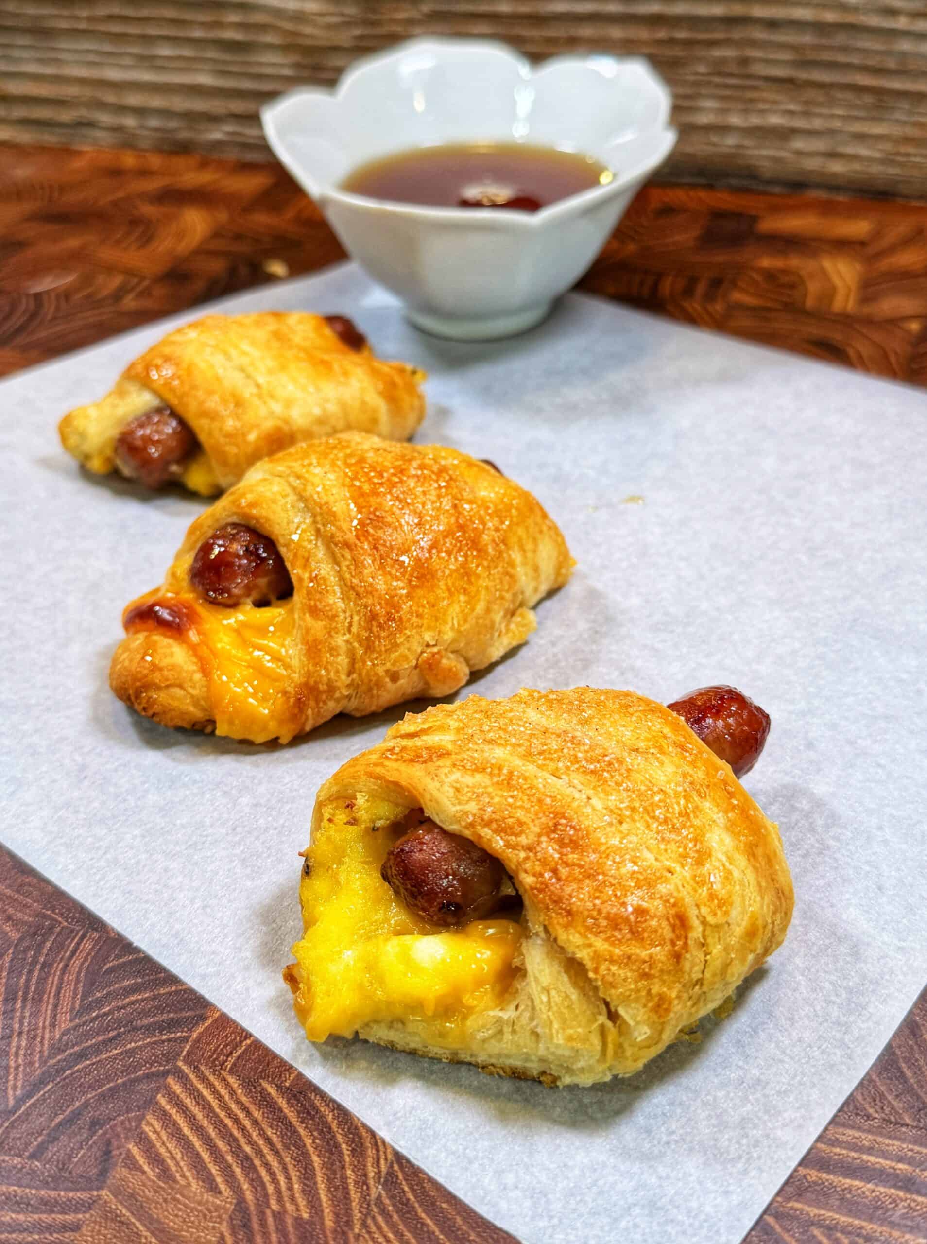 Three sausage, egg, and cheese crescent roll-ups on parchment paper with a small bowl of dipping sauce in the background.