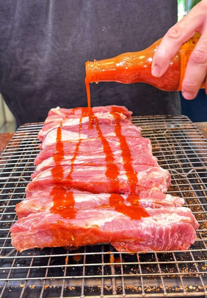 Hot sauce being poured over raw pork ribs on a metal rack.