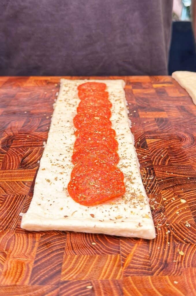 A strip of pizza dough topped with pepperoni slices and Italian seasoning.