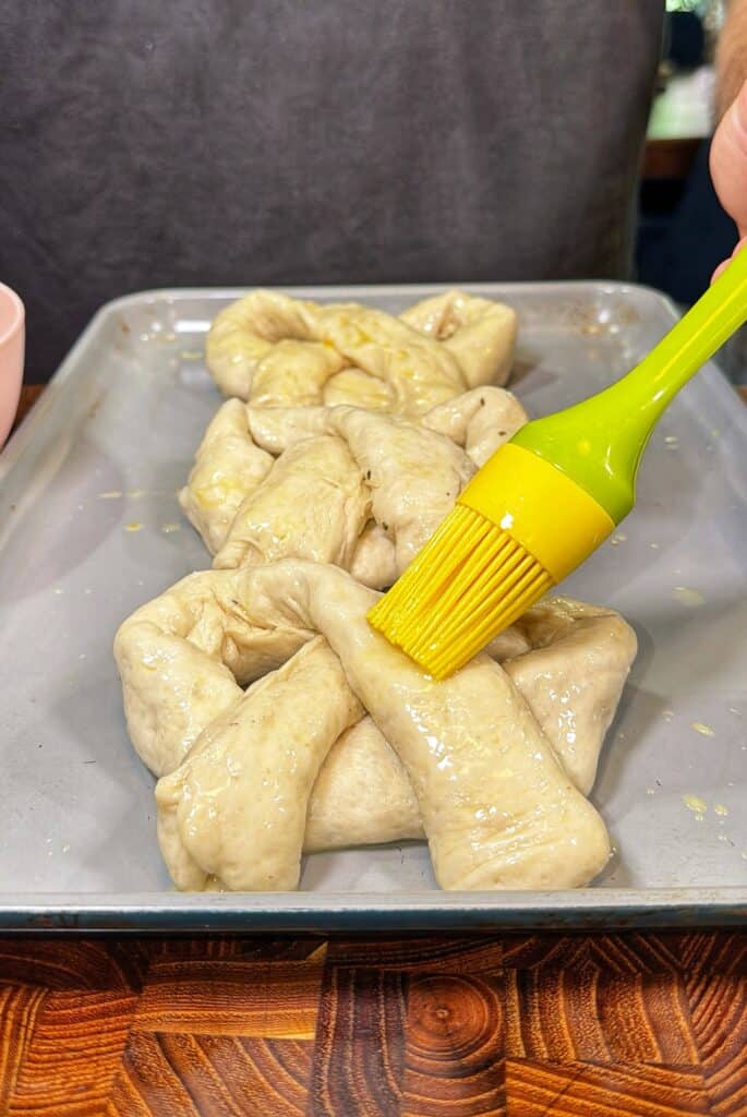 A hand brushes egg wash onto unbaked twisted pretzel dough on a baking sheet.