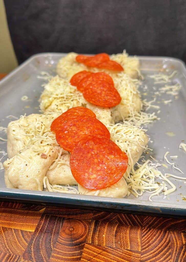 Unbaked stuffed pretzels topped with shredded cheese and pepperoni on a baking sheet.