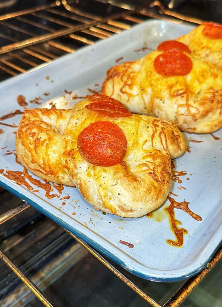 Two stuffed pretzels topped with melted cheese and pepperoni baking on a metal tray in the oven.