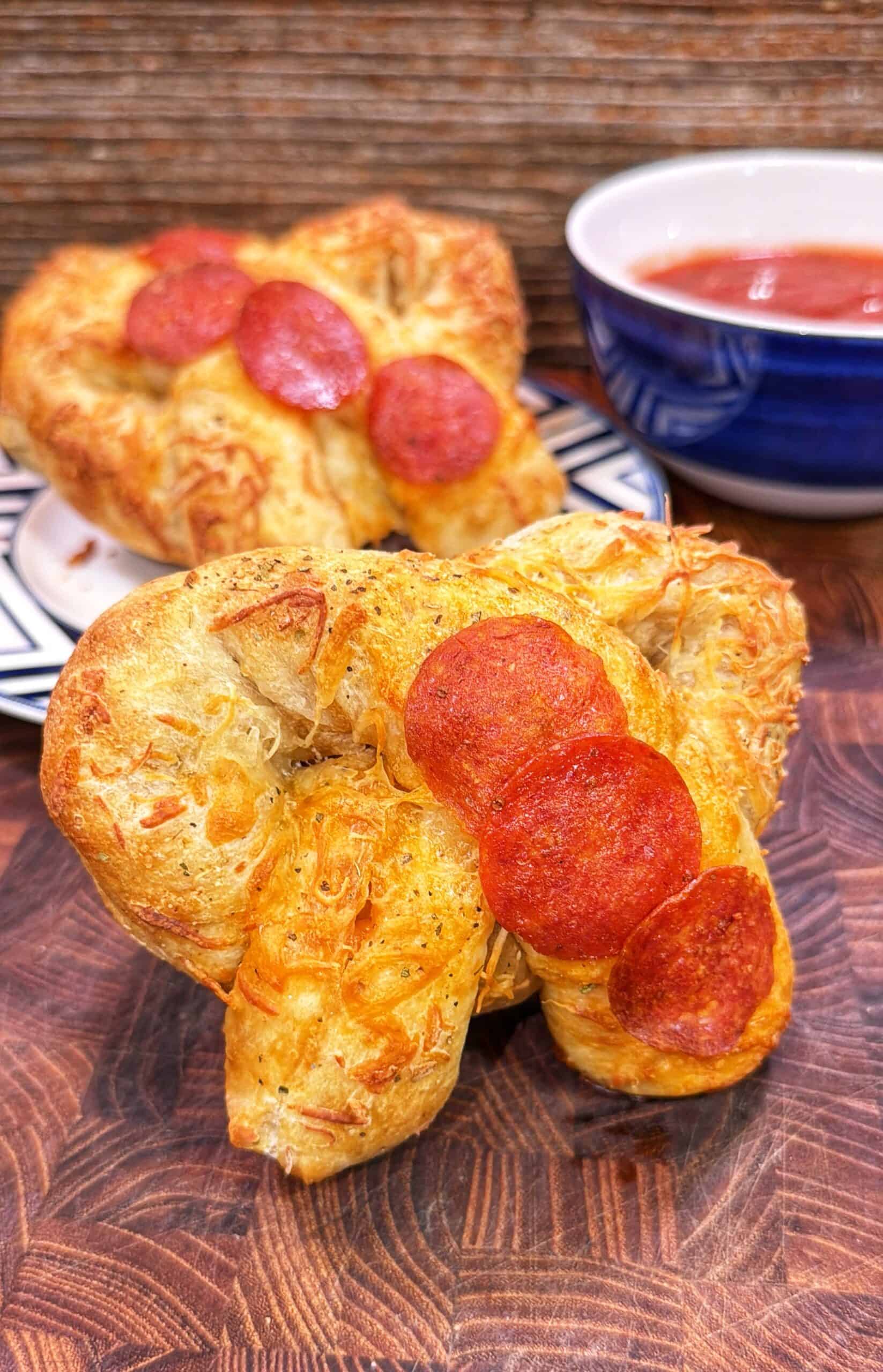 Heart-shaped pepperoni pizza stuffed pretzel with pepperoni slices, with marinara sauce in the background.