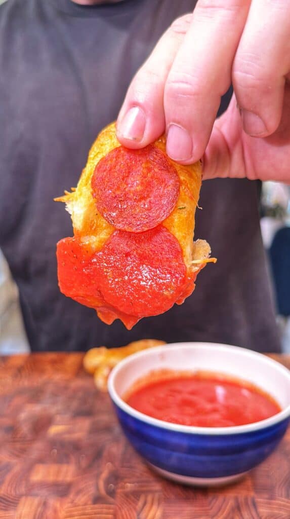 A hand dips a cheesy pepperoni pizza stuffed pretzel into marinara sauce.