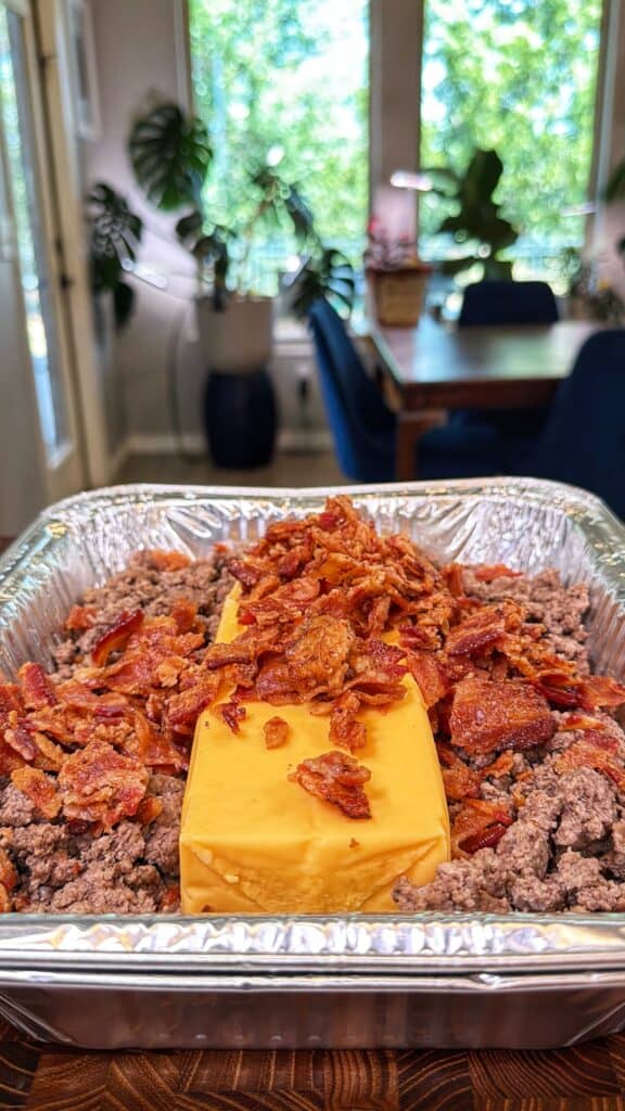 Foil pan of cooked ground beef topped with a block of cheese and crumbled bacon