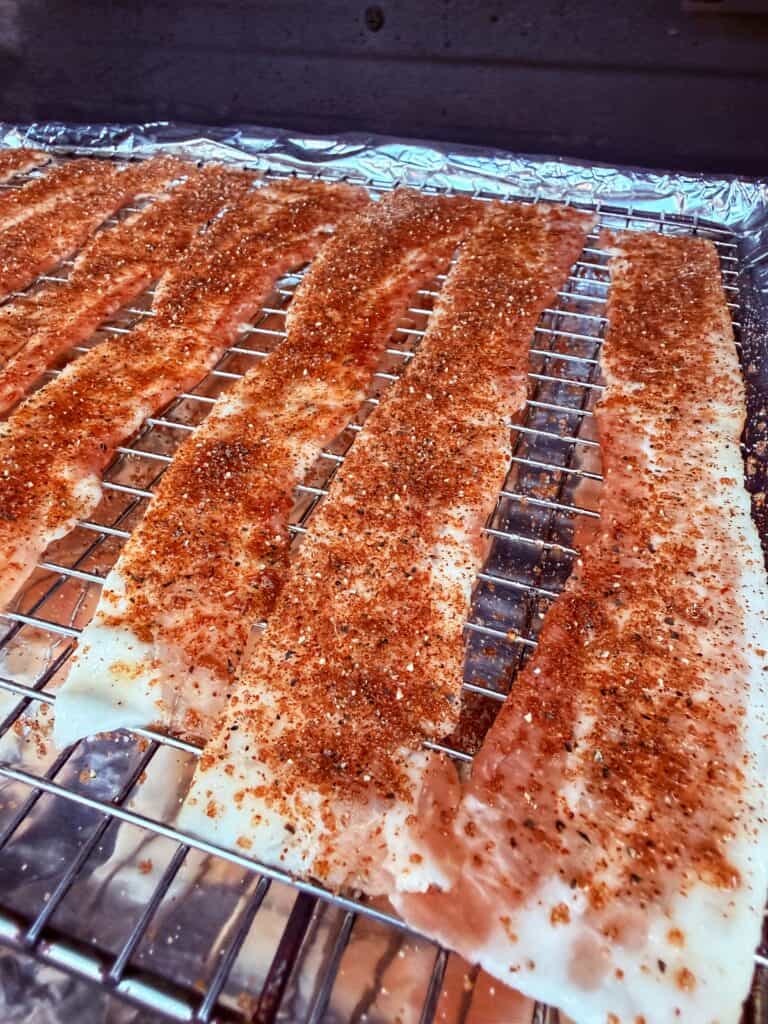 Uncooked bacon strips coated with Nashville hot seasoning are arranged on a wire rack over foil.
