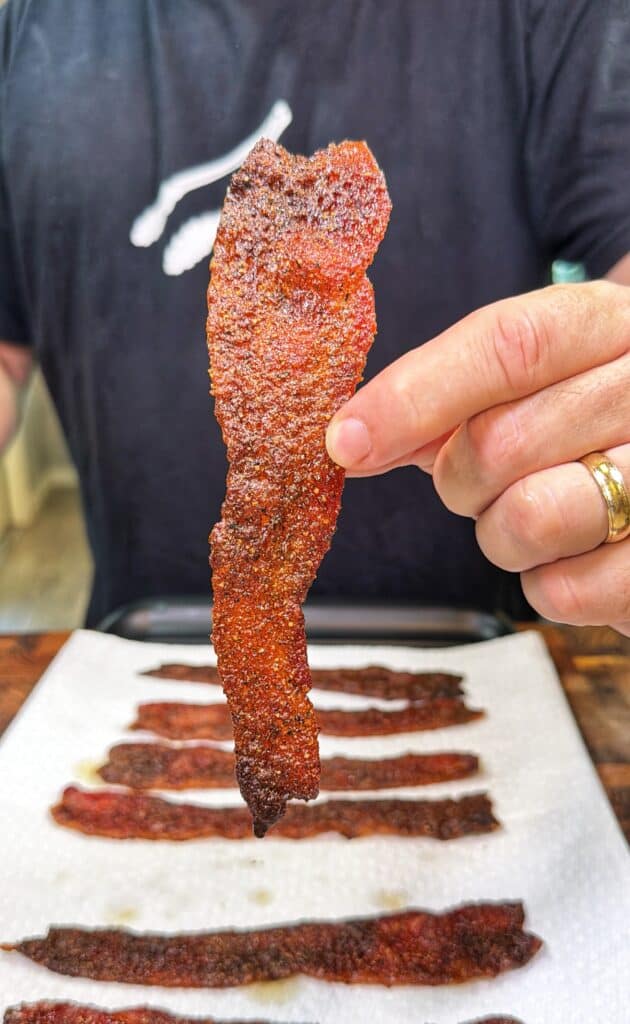 A person holds a strip of Nashville hot candied bacon above several pieces on a paper towel.