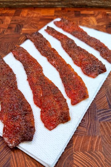 Six strips of Nashville hot candied bacon rest on a paper towel on a wooden cutting board.