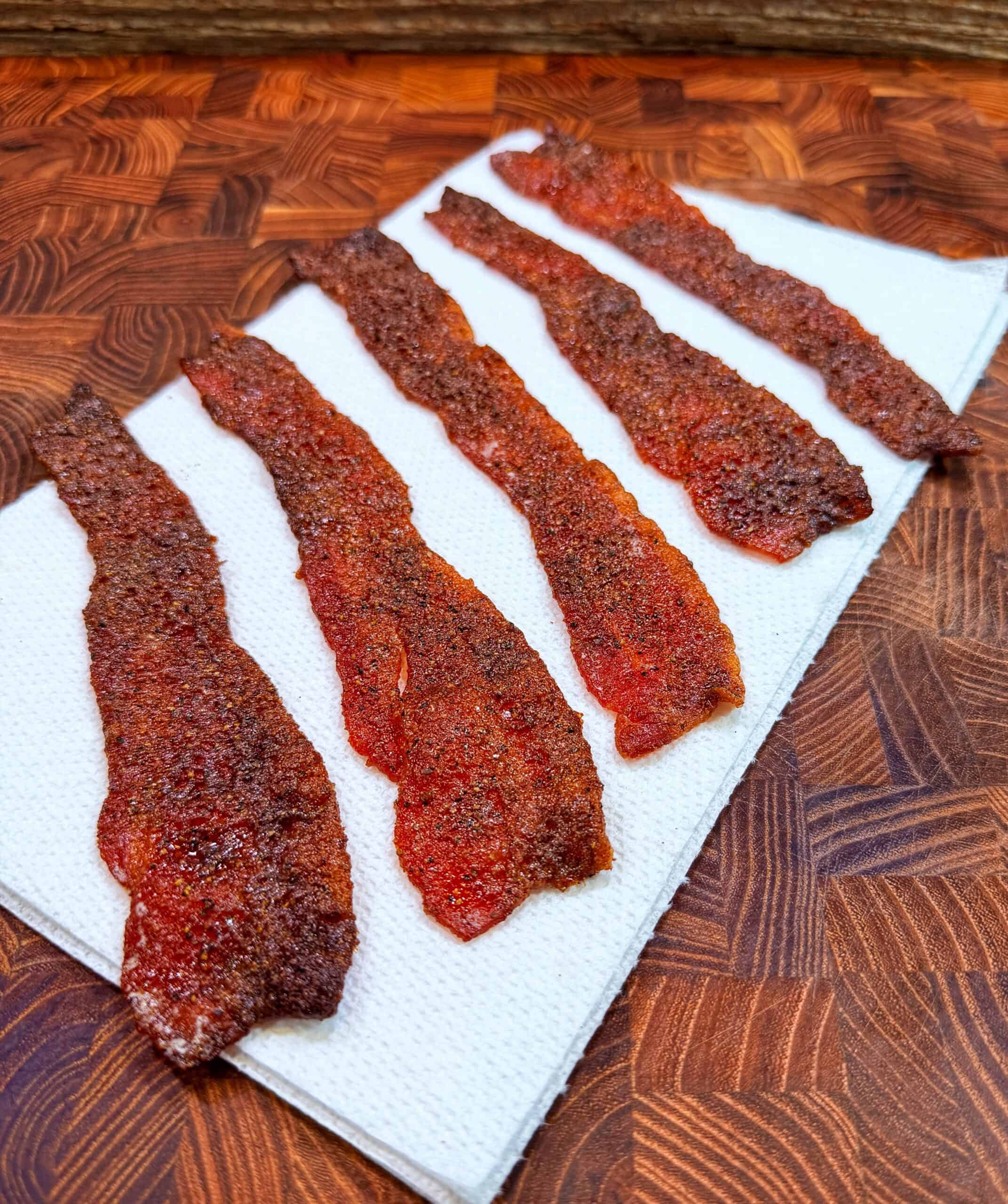 Six strips of Nashville hot candied bacon rest on a paper towel on a wooden cutting board.