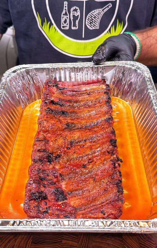 Gloved hand holding a rack of Honey Buffalo ribs over a tray of sauce