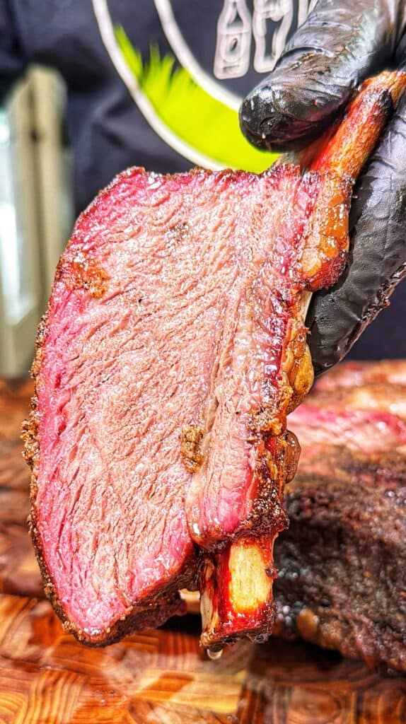 A gloved hand holds a slice of smoked beef rib with bark and a smoke ring above a wooden cutting board