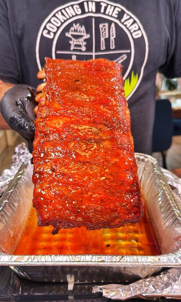 Gloved hand holding a saucy rack of Honey Buffalo ribs over a foil pan of sauce