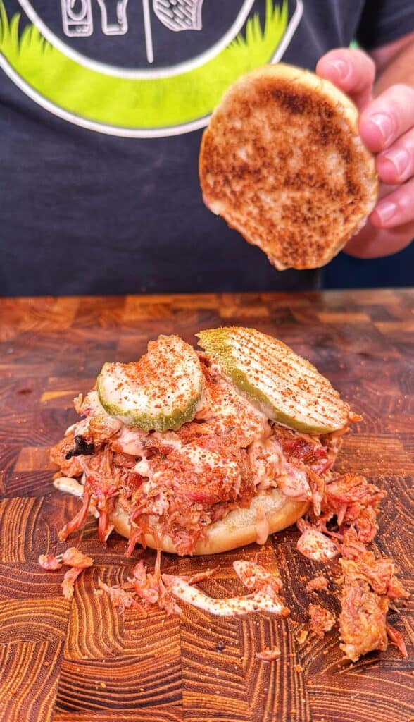 Hand lifting the top bun from a Honey Buffalo shredded rib sandwich with pickles