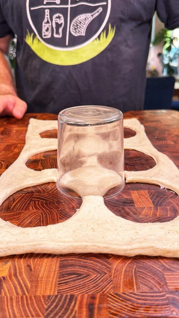 A person uses an upside-down glass to cut rounds from rolled-out pizza dough.