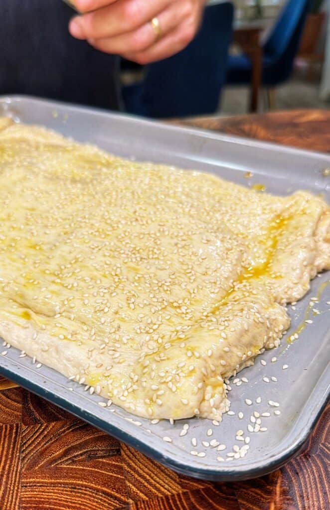 Hand sprinkling sesame seeds over dough on a baking tray.
