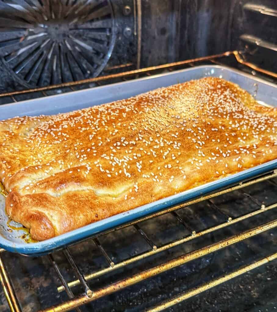 Sesame seed-topped pizza crust baking in a rectangular pan in the oven.