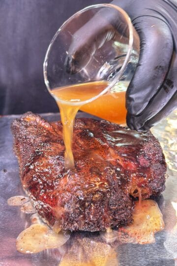 A gloved hand pours liquid from a glass over a seasoned cooked chuck roast in a foil boat.