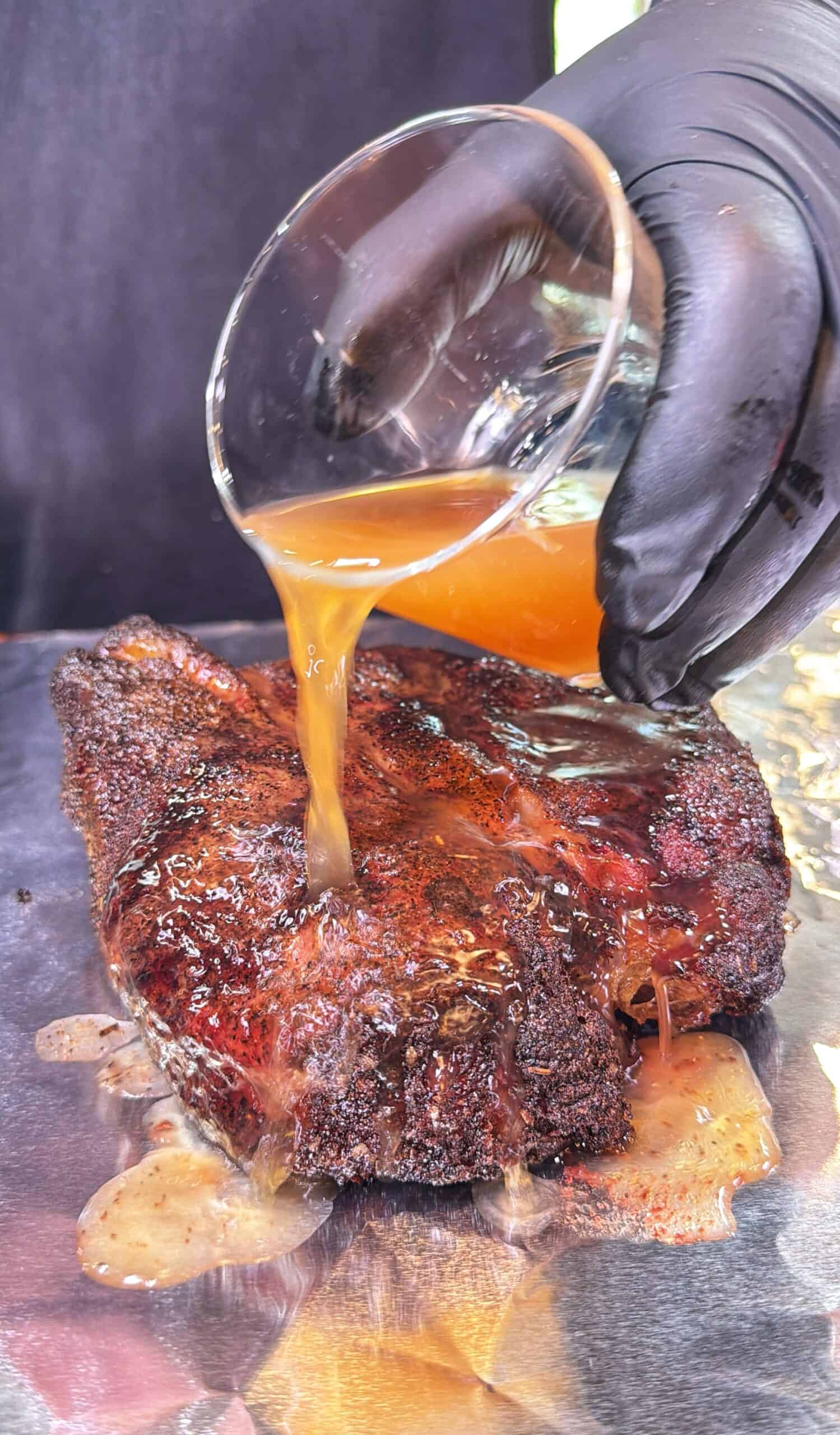 A gloved hand pours liquid from a glass over a seasoned cooked chuck roast in a foil boat.