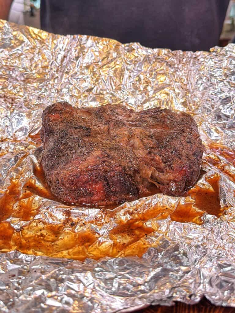 A cooked chuck roast rests in a foil boat with juices pooling around it.