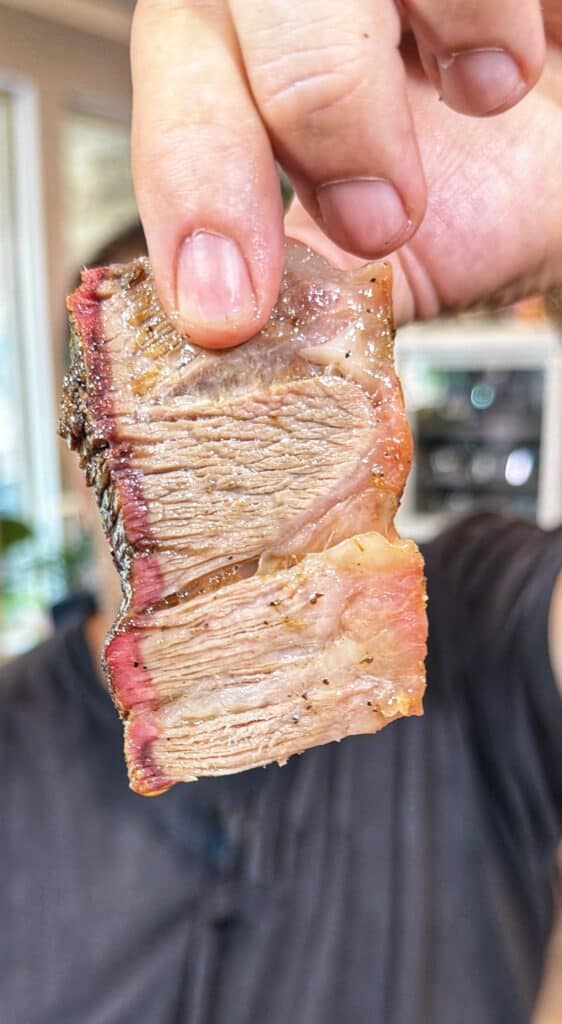 A hand holds a thick slice of smoked chuck roast showing a smoke ring and bark.