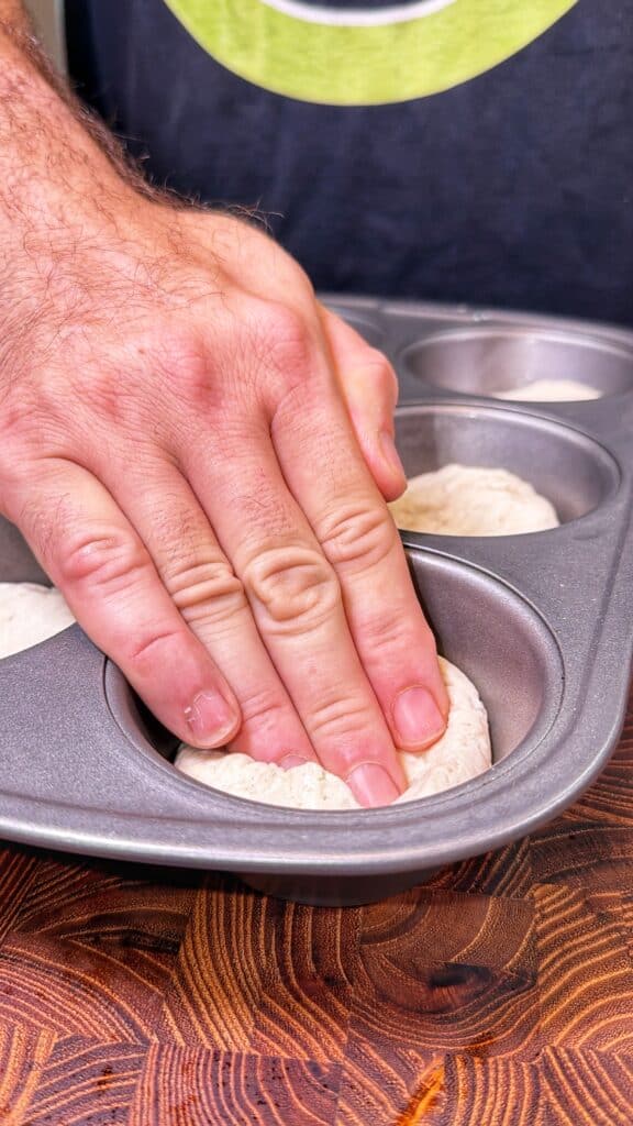 A hand presses biscuit dough into a muffin tin cup.